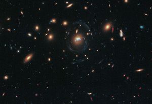 Black background dotted with galaxies. Two reddish galaxies at image center as two dots next to each other, with a thin bluish-white ring curving almost completely around it. Directly below the two galaxies, the filaments appear as a squiggly blue line.